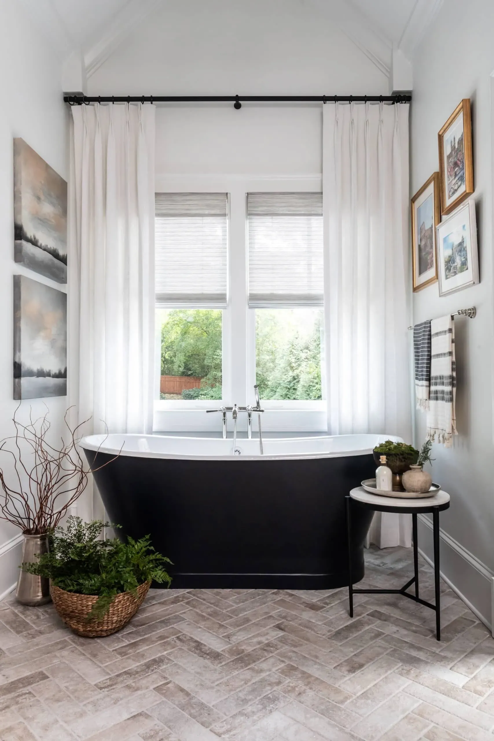 White pinch-pleat curtains on a black rod, layered over cellular shades and flanking a window above a freestanding black tub in a bright bathroom.